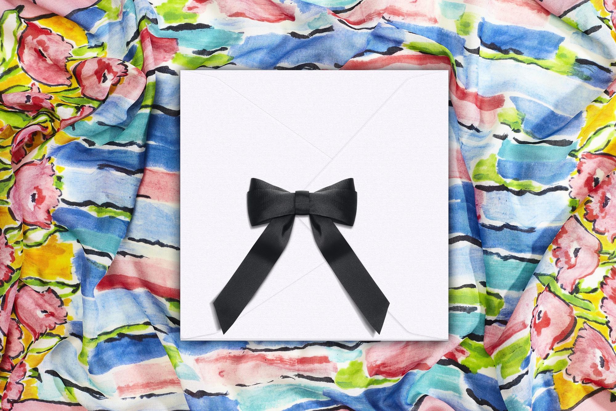 A white By Biehle scarf envelope with a black ribbon bow, lying on a printed silk scarf